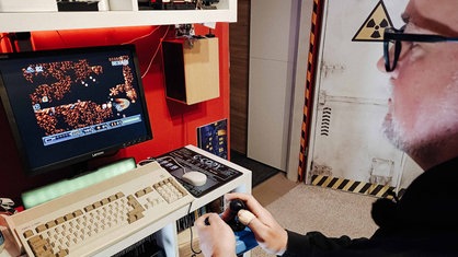 Auf einem kleinen Bildschirm sieht man ein Computerspiel. Tino Menzner steht am Rande des Bildes und hält einen Joystick.