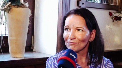 Eine Frau im Interview. Sie hat braune Haare und trägt eine hellblau-weiße Bluse.
