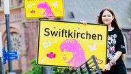 Die Schülerin Aleshanee Westhoff zeigt das Ortsschild "Swiftkirchen" zu Ehren der Musikerin Taylor Swift auf dem Heinrich-König-Platz in Gelsenkirchen in die Kamera.