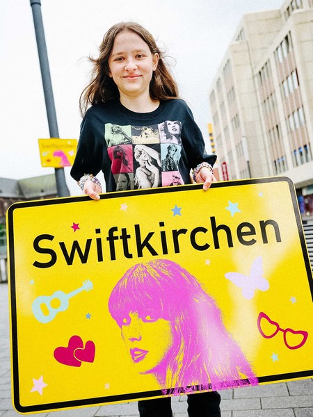 Die Schülerin Aleshanee Westhoff zeigt das Ortsschild "Swiftkirchen" zu Ehren der Musikerin Taylor Swift auf dem Heinrich-König-Platz in Gelsenkirchen in die Kamera.