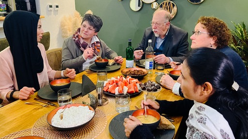 Einige Personen unterschiedlichen Alters sitzen an einem Tisch. Auf dem Tisch befindet sich Essen
