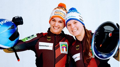 Neele Schuten und Lisa Buckwitz, zwei Frauen mit Mütze und Traininskleidung, lächeln und halten ihre Bob-Helme hoch.