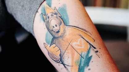 Ein Unterarm einer Frau, auf dem ein buntes "Alf"-Tattoo abgebildet ist