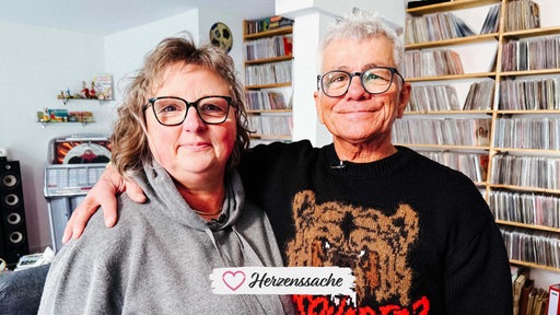Eine Frau und ein Mann mit Brille halten sich im Arm und lächeln in die Kamera.