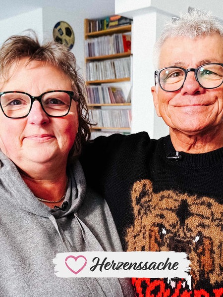 Eine Frau und ein Mann mit Brille halten sich im Arm und lächeln in die Kamera.