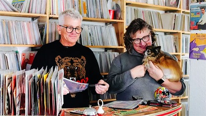 Im Vordergrund stehen ein Mann und eine Frau mit kleinem Hund auf dem Arm an einem höheren Tisch. Im Hintergrund ist eine riesiges Regal voller Schallplatten und CDs.