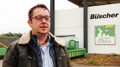  Thomas Overbeeke, ein Mann mit Brille und brauner Lederjacke. Im Hintergrund ist eine Wand mit der Aufschrift "Büscher" und einem Plakat mit der Aufschrift "Frohe Ostern".