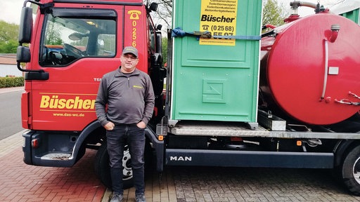 Berni Bösing, ein Mann mit Brille und Kappe steht vor einem roten Reinigungsfahrzeug für Toiletten. Auf dem Fahrzeug ist eine grüne Toilettenkabine mit der Aufschrift "Büscher".