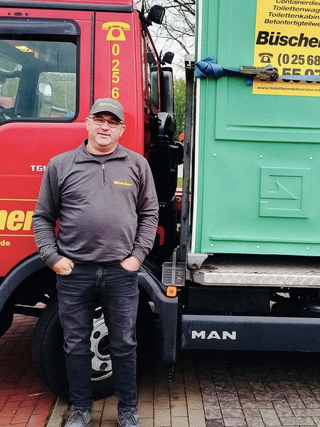 Berni Bösing, ein Mann mit Brille und Kappe steht vor einem roten Reinigungsfahrzeug für Toiletten. Auf dem Fahrzeug ist eine grüne Toilettenkabine mit der Aufschrift "Büscher".