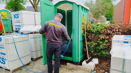 Ein Mann reinigt eine Toilettenkabine mit einem Schlauch. Auf seinem Oberteil ist die Aufschrift "Büscher".