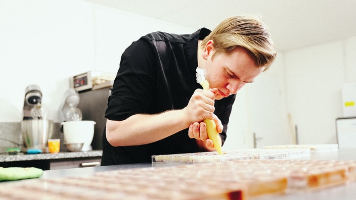 Nils Thiede, ein junger blondhaariger Mann im schwarzen Polohemd, hält eine gefüllte Spitztüte in der Hand und verziert Pralinen.