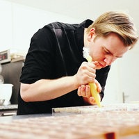 Nils Thiede, ein junger blondhaariger Mann im schwarzen Polohemd, hält eine gefüllte Spitztüte in der Hand und verziert Pralinen.