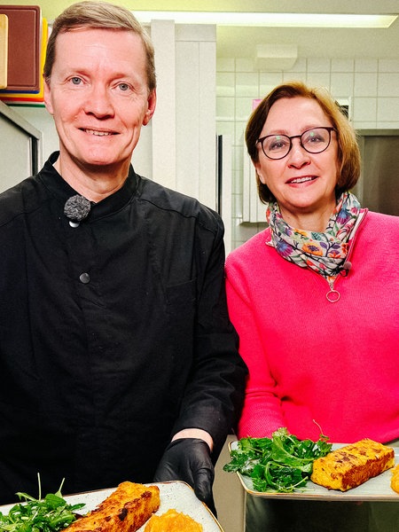 Jürgen Focks und Sabine Behrenbeck stehen in einer Küche und halten auf einem Teller angerichtete Speisen in die Kamera.