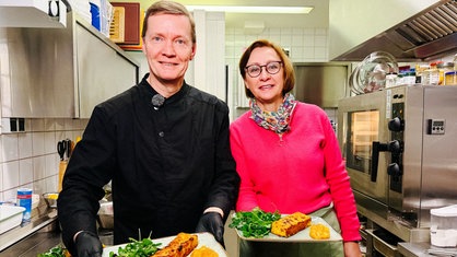 Jürgen Focks und Sabine Behrenbeck stehen in einer Küche und halten auf einem Teller angerichtete Speisen in die Kamera.