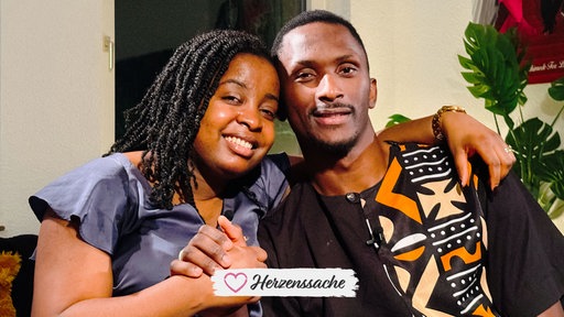 Michèle Leutcha Tchatchou, eine junge Frau in einer violetten Bluse und Archimede Mbazou Kouadjio, ein junger Mann in einem braunen gemusterten Hemd, sitzen innig umarmt nebeneinander und halten sich die Hände.