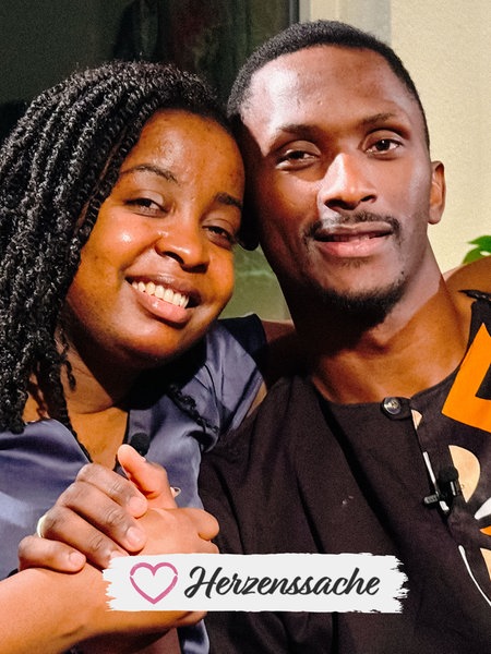 Michèle Leutcha Tchatchou, eine junge Frau in einer violetten Bluse und Archimede Mbazou Kouadjio, ein junger Mann in einem braunen gemusterten Hemd, sitzen innig umarmt nebeneinander und halten sich die Hände.