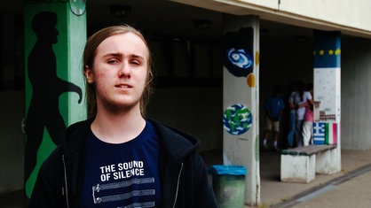Jonathan Bork im Portrait, auf einem Schulhof. Im Hintergrund einige bemalte Pfeiler, die ein Schulgebäude stützen.