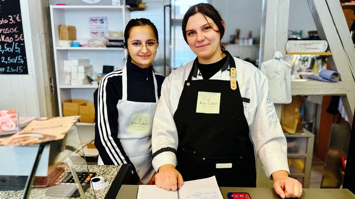 Zwei dunkelhaarige Frauen, Van Reger Shawkat und Krystyna Us, stehen hinter einem Verkaufstresen und halten ein Notizbuch mit handschriftlichen Bestellungen.