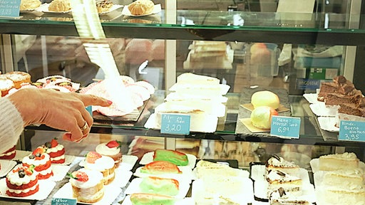 In einer Glasvitrine liegen verschiedene Kuchenstücke und süßes Gebäck, links im Bild zeigt die Hand einer älteren Dame auf eine Reihe mit Zitronenkuchen.