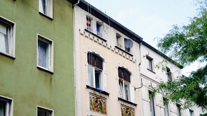 Die Obergeschosse von drei Häusern von außen, schwache Farben und Patina auf den Fassaden, die Fenster sind verschmutzt, Kabel hängen lose die Wand herunter.