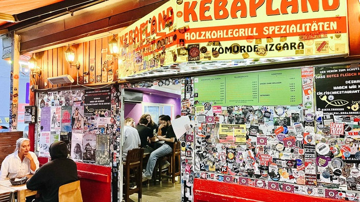 Eine Fassade mit dem Schild "Kebapland". Sie ist vollgeklebt mit Stickern. Draußen und drinnen sitzen Kunden an kleinen Tischen.