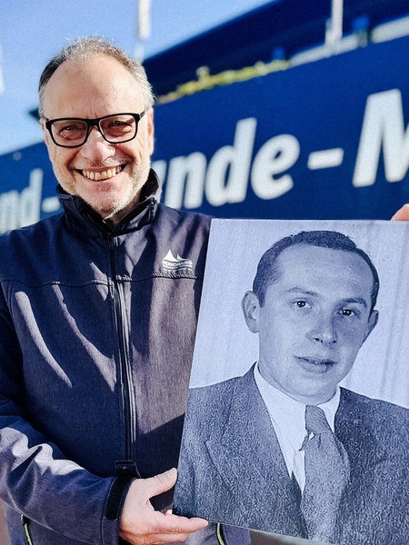 Alfred Brechner steht vor der Tausend Freunde Mauer am Schalker Stadion. Er hält ein Foto von Ernst Alexander hoch und lächelt.
