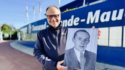 Alfred Brechner steht vor der Tausend Freunde Mauer am Schalker Stadion. Er hält ein Foto von Ernst Alexander hoch und lächelt.