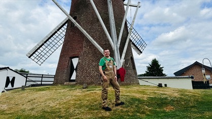 Der Müller Leon Joachims steht in brauner Arbeitskleidung vor seiner historischen Windmühle.