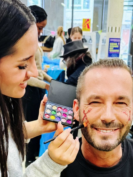 Eine junge Frau hat eine bunte Schminkpalette in der Hand und malt einem Mann etwas auf die Wange