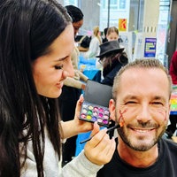 Eine junge Frau hat eine bunte Schminkpalette in der Hand und malt einem Mann etwas auf die Wange