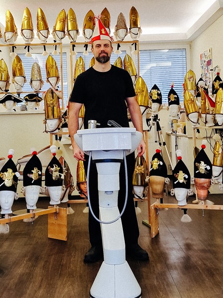 Markus Gurok in einem Friseursalon. Um ihn herum dutzende Puppen mit Karnevalsschmuck.