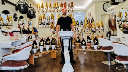 Markus Gurok in einem Friseursalon. Um ihn herum dutzende Puppen mit Karnevalsschmuck.