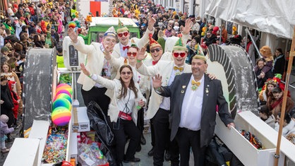 Auf einem großen Wagen stehen Männer udn Frauen in weißen Anzugjacken, die lächeln und winken, rechts und links von dem Karnevalswagen stehen verkleidete Menschen.