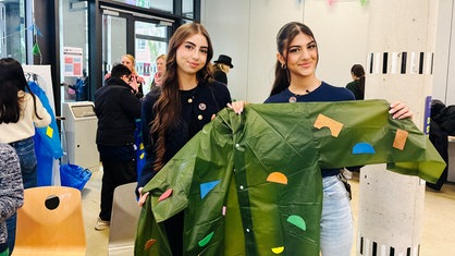 Zwei junge Frauen stehen nebeneinander und halten einen dunkelgrünen Plastikmantel mit aufgeklebten bunten Emblemen