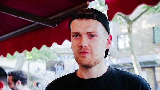 Junger Mann mit schwarzer Cap, schwarzem T-Shirt und Drei-Tage-Bart, hinter ihm sieht man Ausläufer einer roten Markise, unter der er steht