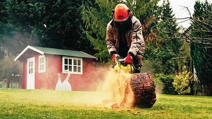 Ein junger Mann in einer beigen Jacke, orangem Schutzhelm und Schutzhandschuhen, beugt sich mit einer gelben Kettensäge über einen Holzstamm und sägt.