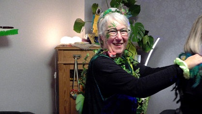 Organisatorin Ute Drießen, eine Frau mit kurzen, weißen Haaren mit künstlichen Pflanzen als Dekoration im Haar und um den Oberkörper gewickelt, läuft in einer Polonaise und lacht in die Kamera.