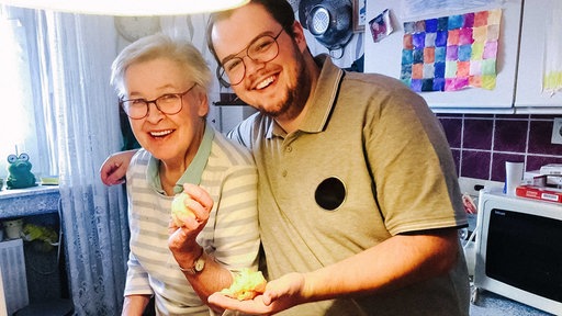 Melvin Flor legt seinen Arm um seine Großmutter. Sie stehen in der Küche und lachen.