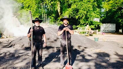 Die Köhler Andre Verheyen und Tim Petermann stehen vor dem qualmenden Kohlemeiler.