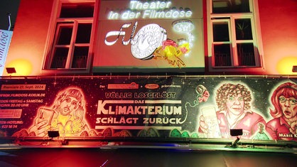 Das "Theater in der Filmdose" in Köln von außen, mit Schriftzug und einem großen Plakat, das das aktuelle Stück "Völlig losgelöst - Das Klimakterium schlägt zurück" ankündigt