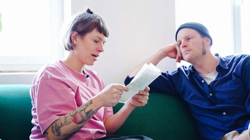 Eine Frau mit kinnlangen,braunen Haaren und rosa Pullover sitzt auf einer Couch und liest einen Brief vor. Neben ihr sitzt ein Mann mit Mütze und blauem Hemd und hört ihr zu.