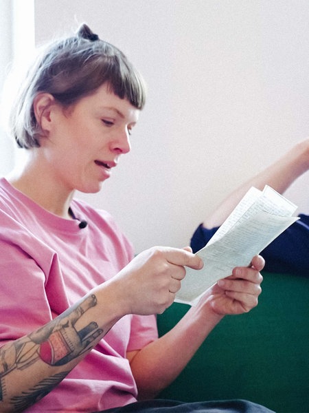 Eine Frau mit kinnlangen,braunen Haaren und rosa Pullover sitzt auf einer Couch und liest einen Brief vor. Neben ihr sitzt ein Mann mit Mütze und blauem Hemd und hört ihr zu.