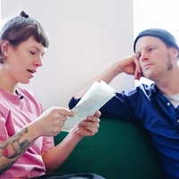 Eine Frau mit kinnlangen,braunen Haaren und rosa Pullover sitzt auf einer Couch und liest einen Brief vor. Neben ihr sitzt ein Mann mit Mütze und blauem Hemd und hört ihr zu.