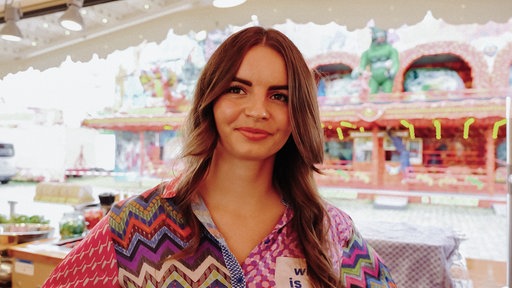 Lisa Lemoine steht in der Bude ihrer Familie, im Hintergrund ein buntes Kirmes-Fahrgeschäft