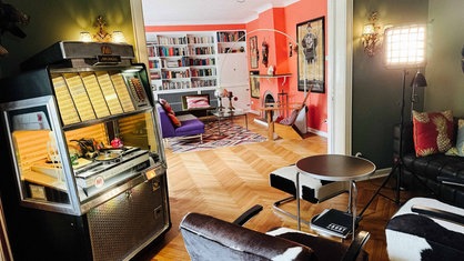 Dunkelgrünes Wohnzimmer mit Jukebox. Hinter einem Durchgang die andere Hälfte des Wohnraumes, in orange gestrichen.