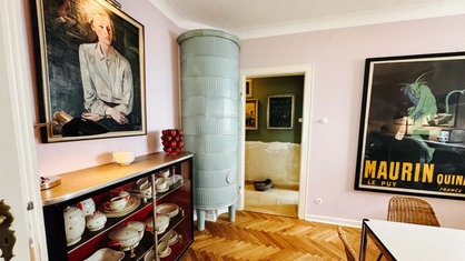 Ein Wohnraum in rosa gestrichen. Links steht eine Vitrine mit altem Geschirr. Große Bilder hängen an den Wänden.