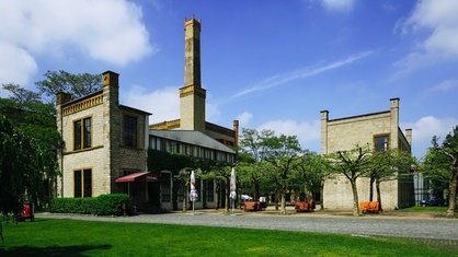 Ehemaliges Industriegebäude aus beigem Stein, davor Bäume und Gastronomie