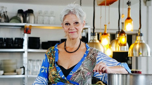 Angela von der Goltz sitzt in ihrer Manufaktur. Mit ihrem linken Arm stützt sie sich gelassen auf einen großen silbernen Topf.