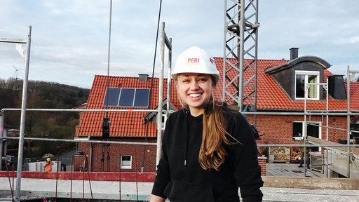 Eine junge Maurermeisterin steht mit Helm auf einer Baustelle.