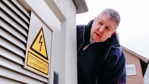 Eine Frau blickt auf einen Stromverteiler, darauf steht das Schild "Hochspannung Lebensgefahr".